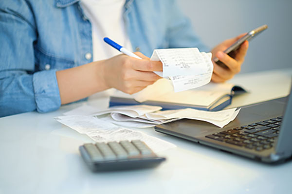 A person surrounded by a bunch of receipts, a calculator, laptop, and notebook, calculating finances on their phone.