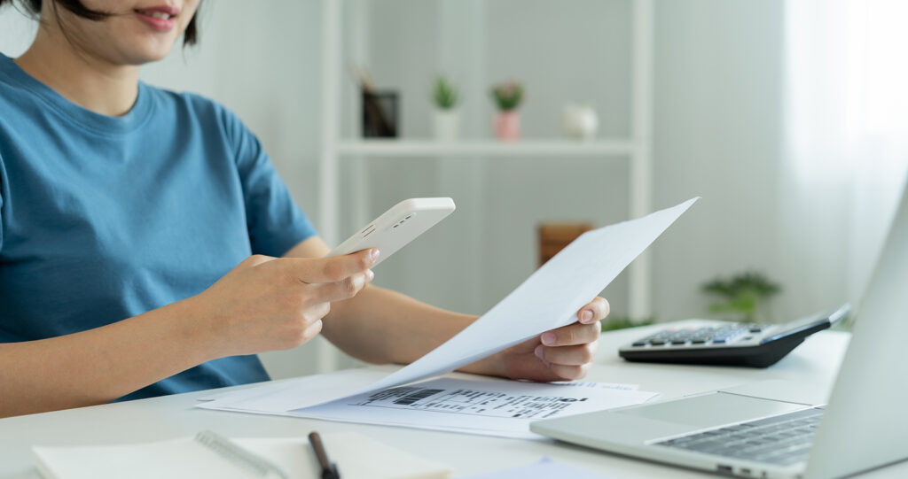 A woman sits at a desk. She's holding a paper in her hands. There's also a laptop open in front of her and a calculator.