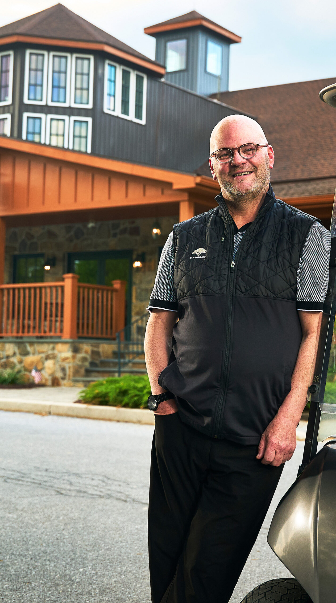 A man is smiling at the camera while leaning to his right on a golf cart. In the background there is the clubhouse building for a golf course.