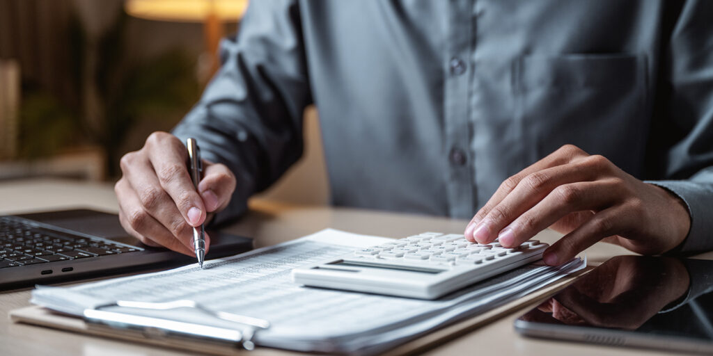 a person sits at a desk with a stack of papers and a calculator. They're holding a pen in their left hand and pointing at the paper like they are reading through the page.