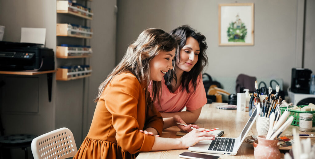 Two women smile while reviewing something on the laptop in front of them. They appear to be in an office space. There is a printer behind them and shelving with items filed away.