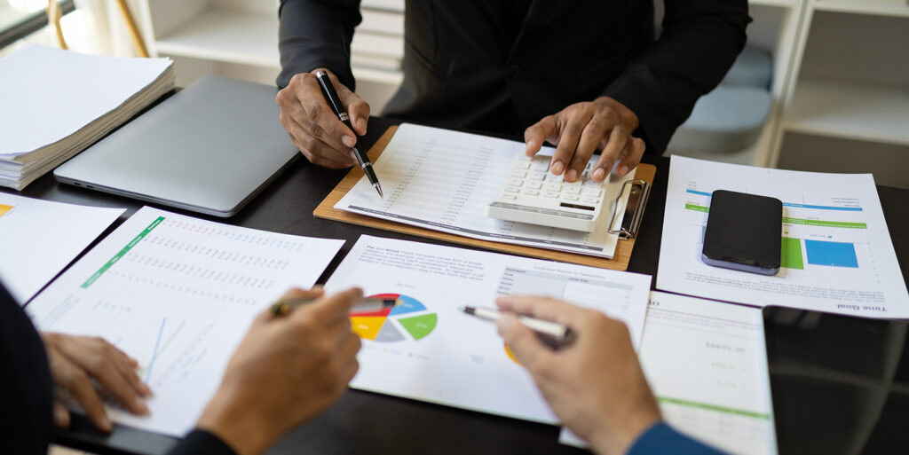 Three set's of hands are reviewing charts and paperwork to represent benchmarking data for businesses.