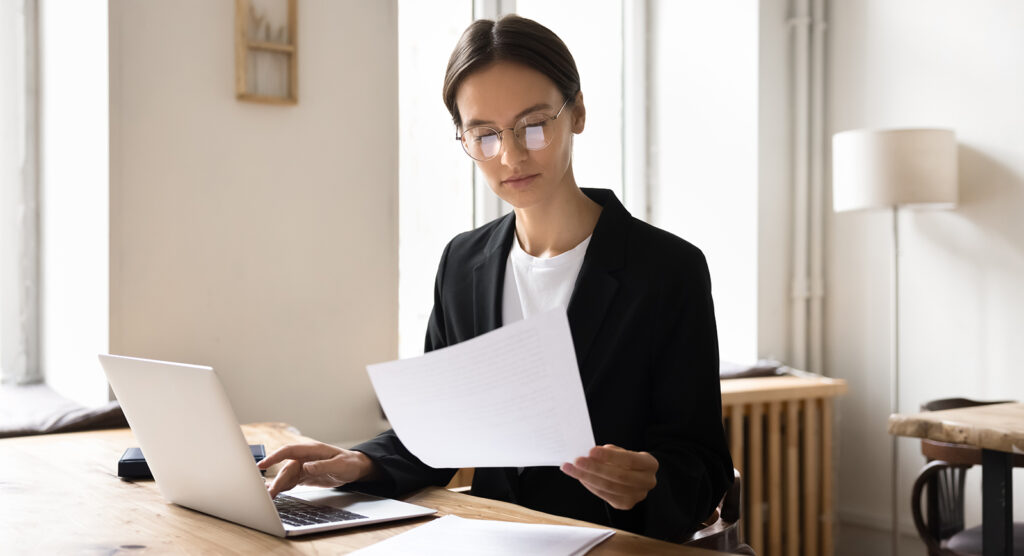 A woman in sits at a desk with a laptop in front of her while holding a sheet of paper. It represents the blog, "The Value of Linking Your Business Bank Account to Your Accounting Software."