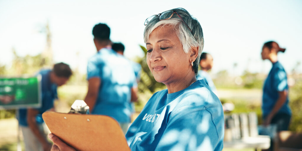 A woman is in the foreground holding a clipboard. She is wearing a blue t-shirt that says, "volunteer." In the background, there are four more people with the same t-shirt on. The picture is to represent non-profits.
